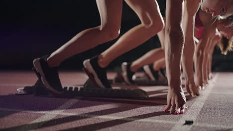 A-row-of-runners-womens-crouch-in-the-starting-position-before-beginning-to-race.-Females-start-with-running-shoes-on-the-stadium-from-the-start-line-in-the-dark-with-spotlights-in-slow-motion.